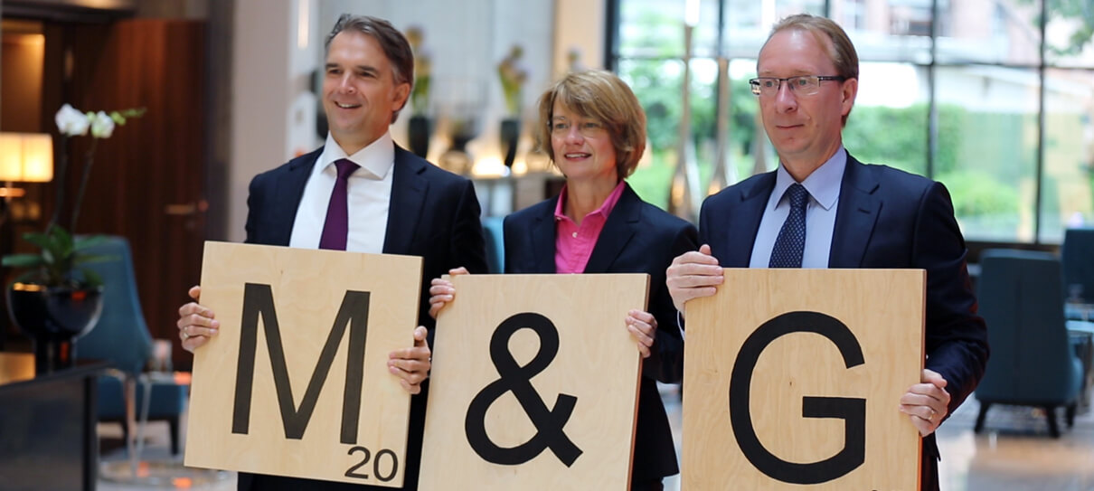 Werner Kolitsch, Richard Woolnough und Sabine Schumann von M&G Investments stehen mit drei Scrabblesteinen in der Hand nebeneinander. Die Buchstaben formen den Firmennamen M&G. Screenshot Eventfilm - Filmproduktion Karlsruhe mp-film für das Unternehmen in Frankfurt von der M&G Jahreskonferenz produziert hat.